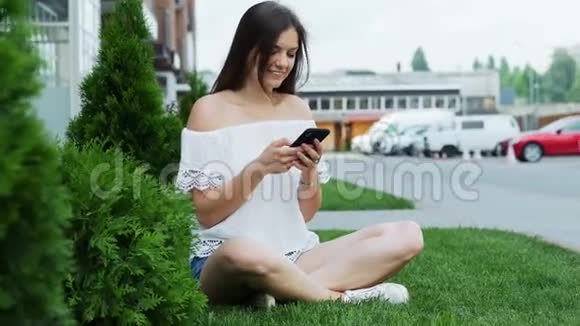 年轻漂亮的女人坐在酒店附近的草地上使用智能手机视频的预览图