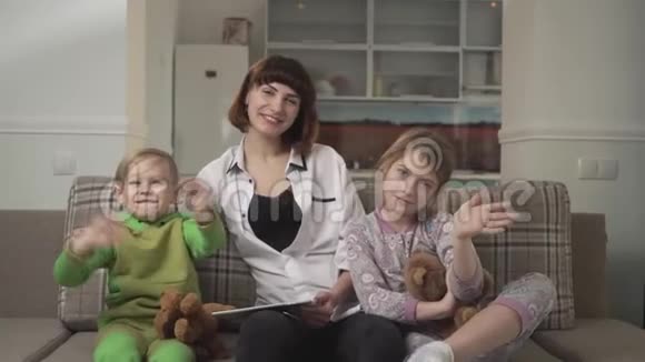 快乐的母亲和两个孩子坐在沙发上向相机挥手家庭假日视频的预览图