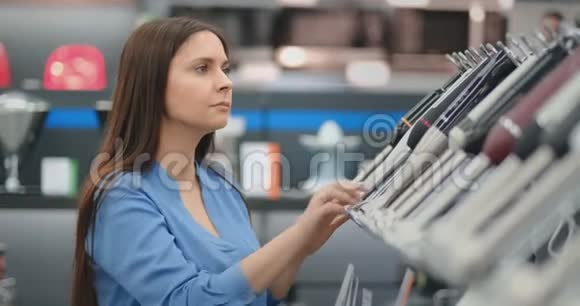 在电器商店里一个穿着衬衫的黑发女人通过观看和拿着设备来选择搅拌机购物视频的预览图