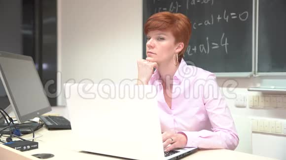 年轻女教师一边浏览文件一边坐在办公桌上手提电脑在现代教室里视频的预览图