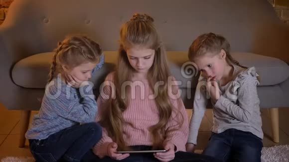 三个漂亮的白种人女孩坐在地板上在舒适的家里专心地看着平板电脑的画像视频的预览图