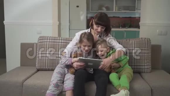 幸福妈妈和两个孩子坐在沙发上看着平板电脑家庭假日视频的预览图