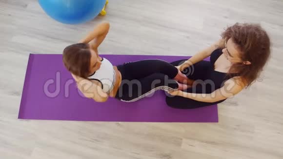 年轻女性在工作室健身一个女人抽着腹肌躺在紫色的瑜伽垫上而另一个女人视频的预览图