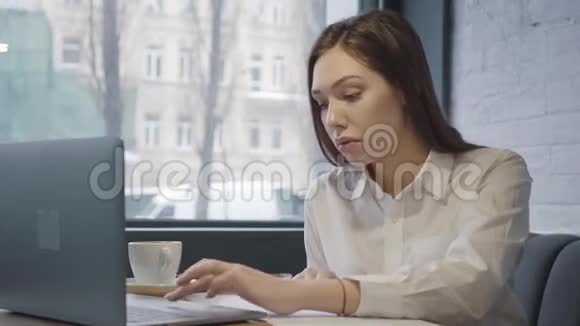 成功的集中女性使用她的笔记本电脑坐在咖啡馆的桌子上坐在靠近窗户的沙发上的女孩视频的预览图