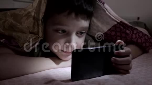一个男孩躺在家里的床上在毯子下面的黑暗中在智能手机上放松和观看视频视频的预览图