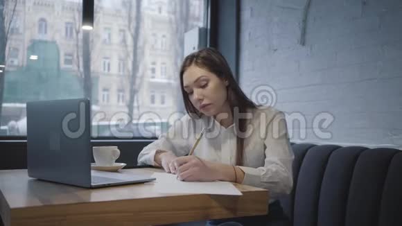 专心致志的女人用她的笔记本电脑坐在咖啡馆的桌子上坐在靠近窗户的沙发上喝咖啡的女孩视频的预览图