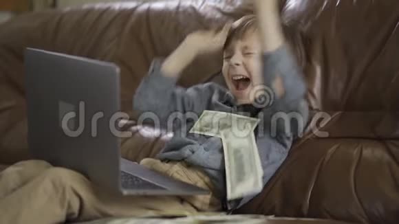 肖像可爱可爱的小男孩坐在皮革沙发上腿上放着笔记本电脑把钱扔在里面视频的预览图