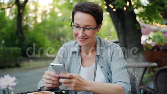 微笑的女士用智能手机触摸屏幕微笑的街外咖啡厅视频的预览图
