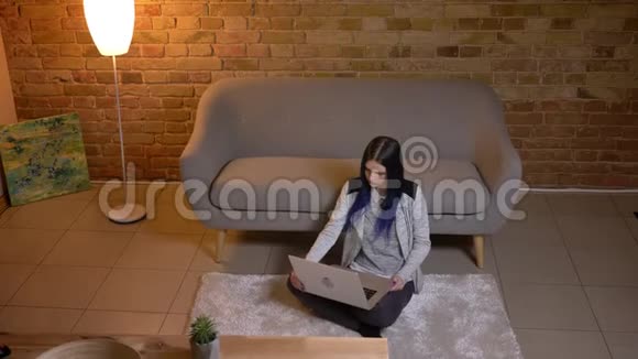 在一间舒适的公寓里年轻漂亮的白种人女性坐在地板上看一部笔记本电脑上的电影视频的预览图