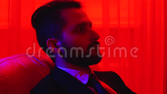 一个穿着商务服满脸胡须的疲惫男人靠在椅子上用明亮的红光遮住眼睛的画像视频的预览图