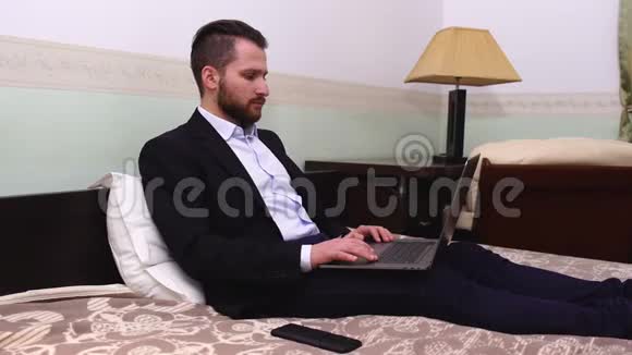 穿西装的英俊商人坐在床上拿着笔记本电脑英俊的胡子男在卧室工作视频的预览图