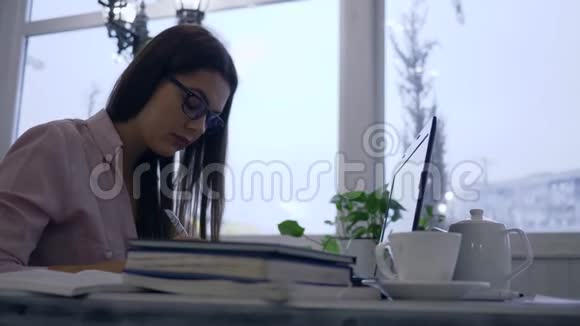 在电子学习过程中聪明的学生女孩使用现代上网本和书籍并在咖啡馆的桌子旁的笔记本上写笔记视频的预览图
