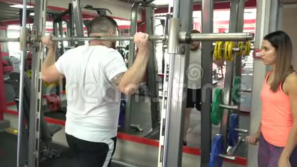 一个留着胡子的男人在健身房里做运动在一个人的监督下他用杠铃蹲着视频的预览图