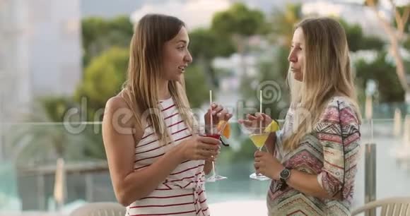 两个有魅力的女人正在游泳池和棕榈树的背景下交谈和喝鸡尾酒视频的预览图
