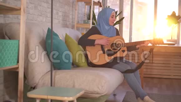 年轻的穆斯林妇女坐在沙发上弹吉他视频的预览图