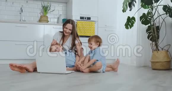 妈妈带着两个小孩坐在一间现代化公寓的地板上明亮的房间里有一台笔记本电脑注意视频的预览图