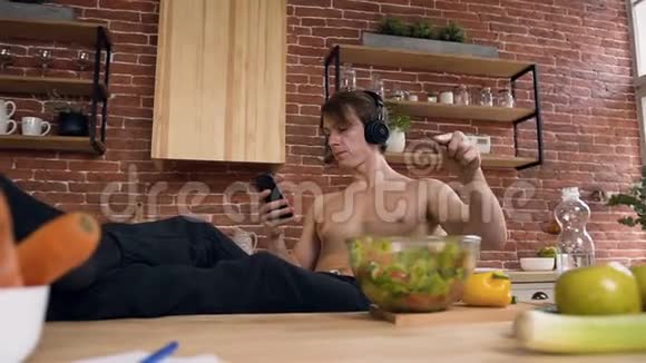 年轻的运动员赤裸的躯干坐在厨房的桌子后面一边吃纯素沙拉一边用耳机听音乐视频的预览图