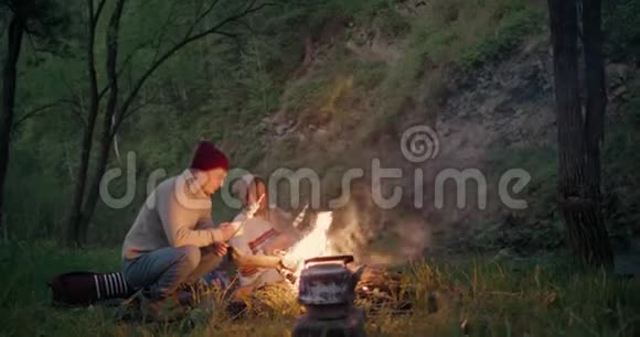 一对年轻夫妇坐在河边的森林里聊天煎棉花糖视频的预览图