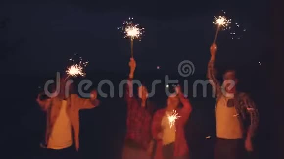 兴奋的年轻人在森林里的火堆旁用孟加拉灯跳舞欢笑着享受音乐聚会视频的预览图