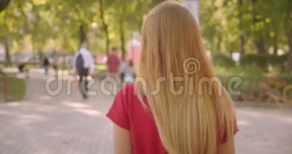 特写后景照片年轻的金发碧眼的长头发欢快的女性在公园里散步转身看镜头视频的预览图