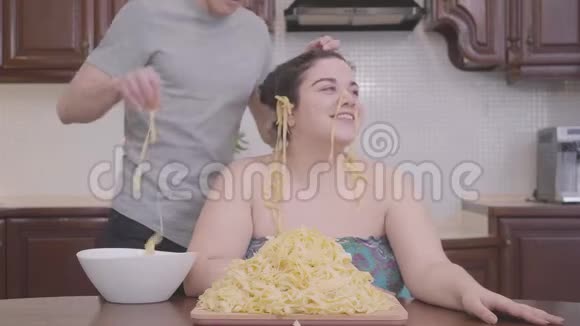 肖像漂亮丰满的女人坐在家里的厨房里那个苗条的金发男人把面条挂在他肥胖的耳朵上视频的预览图