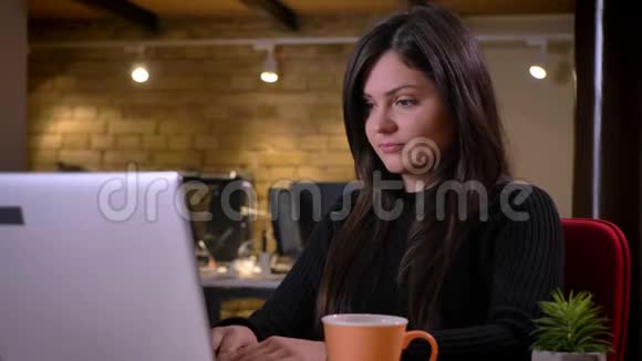 成年白人黑发女商人在笔记本电脑上打字端着杯子喝咖啡的特写照片视频的预览图