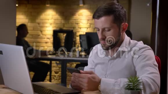 年轻白种人商人用手机和听音乐坐在笔记本电脑前的特写肖像视频的预览图