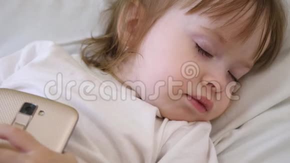 小婴儿手里拿着电话在枕头上睡着了孩子在床上睡觉手里拿着智能手机视频的预览图