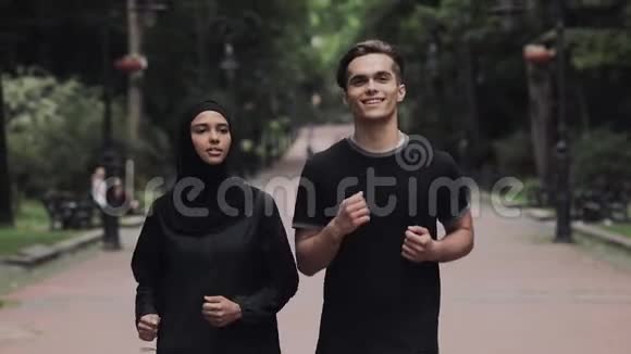 年轻的高加索男人和穿着Hijab的穆斯林女人看起来兴奋和放松的微笑和聊天在公园里跑步视频的预览图