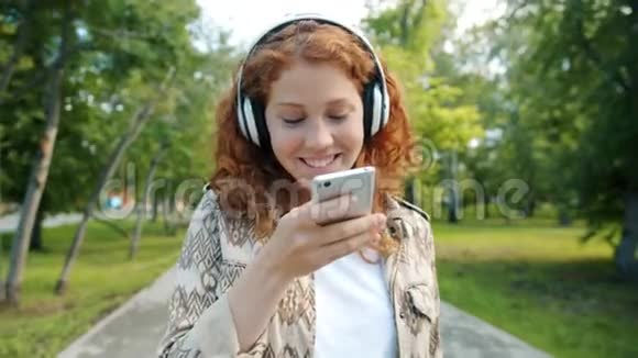 美丽的女孩在绿色公园用智能手机耳机听音乐视频的预览图
