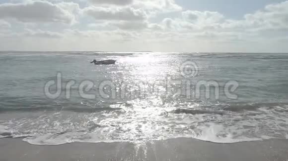 一艘孤立的小船在海面上缓慢移动海浪拍打着海岸阳光反射在背光和多云中视频的预览图