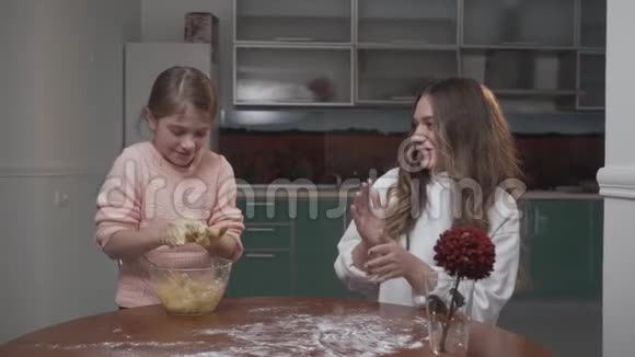 年长和年轻的女孩在厨房的桌子上揉面团两个姐妹坐在桌子旁准备蔬菜沙拉视频的预览图