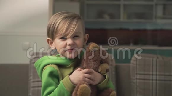 一个穿睡衣的小可爱滑稽男孩抱着一只泰迪熊站在客厅沙发旁边的肖像快乐视频的预览图