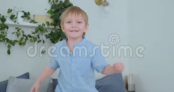 一个长着白色头发和蓝色衬衫的英俊男孩正在沙发上跳跃慢镜头地看着相机视频的预览图