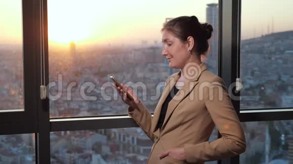 办公室里的年轻女商人正在窗口附近浏览电话可以看到城市全景视频的预览图