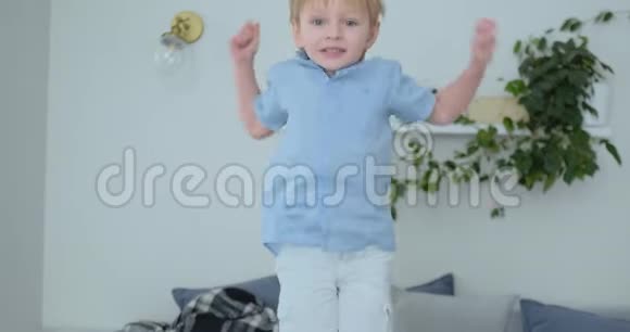 一个长着白色头发和蓝色衬衫的英俊男孩正在沙发上跳跃慢镜头地看着相机视频的预览图