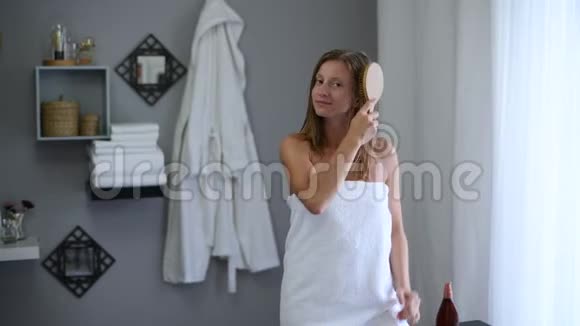 淋浴后裹着毛巾的漂亮女人用木毛刷刷她湿漉漉的头发视频的预览图