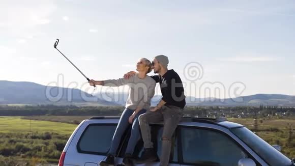 年轻的时髦夫妇在智能手机上自拍坐在山顶背景的汽车屋顶上视频的预览图
