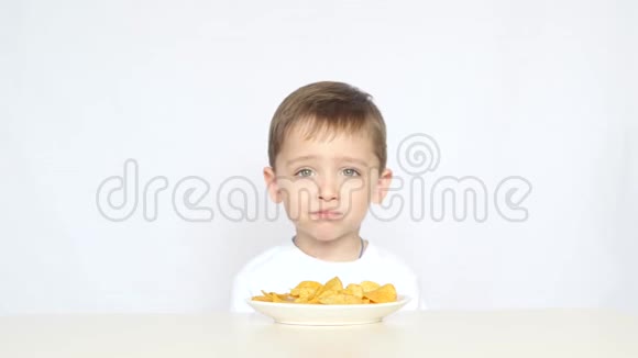 一个可爱的小男孩坐在桌子前的一盘薯片在白色的背景上孩子吃薯片视频的预览图