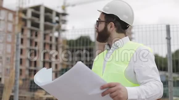 一位成功的年轻工程师或建筑师戴着白色头盔的肖像在他的建筑图纸中寻找视频的预览图