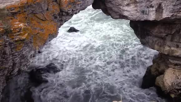 海上风暴巨大的波浪在岩石海岸的石拱下破碎白色泡沫在水面上视频的预览图