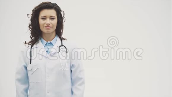幸福的年轻微笑女孩医生的肖像穿着白色长袍在灰色背景上均匀交叉的双手视频的预览图