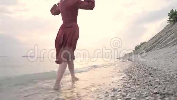 夕阳西下一个穿着红色长裙戴着草帽的年轻漂亮女孩在风和日丽的天气里沿着海滨散步视频的预览图