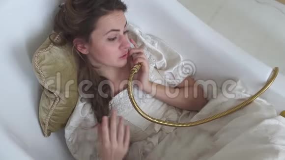穿着球服的有趣的年轻女子躺在浴室里洗澡希望假装是电话在上面聊天视频的预览图