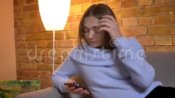 年轻可爱的白种人女性在舒适的沙发上坐着一边打电话发短信一边整理头发特写照片视频的预览图