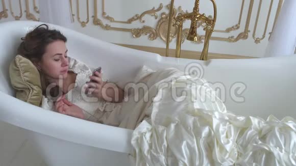漂亮的年轻女孩穿着白色的复古连衣裙躺在空荡荡的浴室里打着手机可爱的女人说话视频的预览图