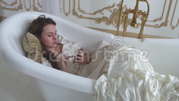 年轻女子穿着球服躺在浴缸里用枕头和手机短信女孩用小工具视频的预览图