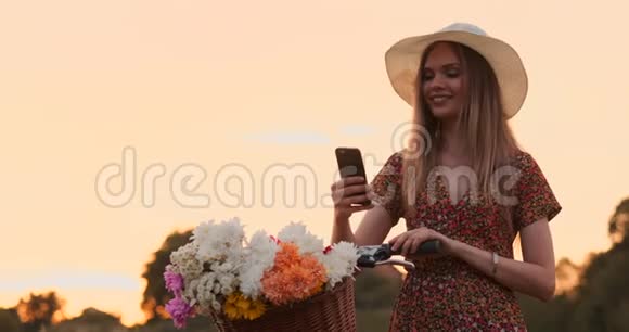 戴着帽子的漂亮金发女郎骑着自行车看着手机屏幕车把上放着花的篮子视频的预览图