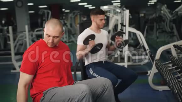 健身房里的两个人坐在长凳上通过举起哑铃来训练二头肌视频的预览图