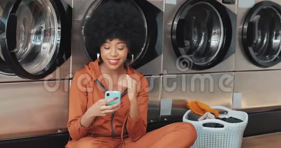 在洗衣店用智能手机微笑的年轻女人非裔美国学生在智能手机上有好消息自我视频的预览图
